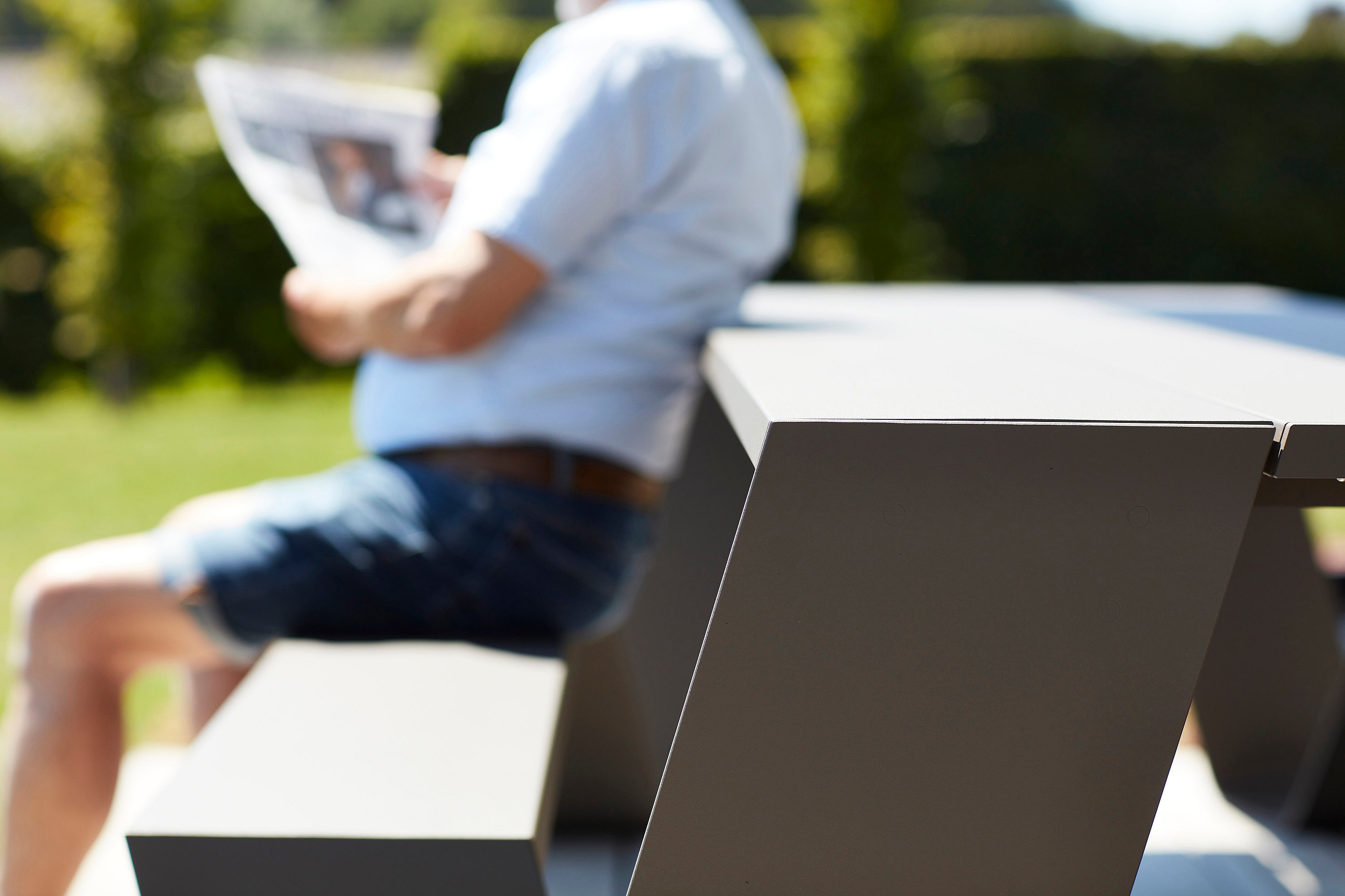 Close-up of the corner of a grey Hopper AA table and bench, with a man reading a newspaper softly blurred in the background.