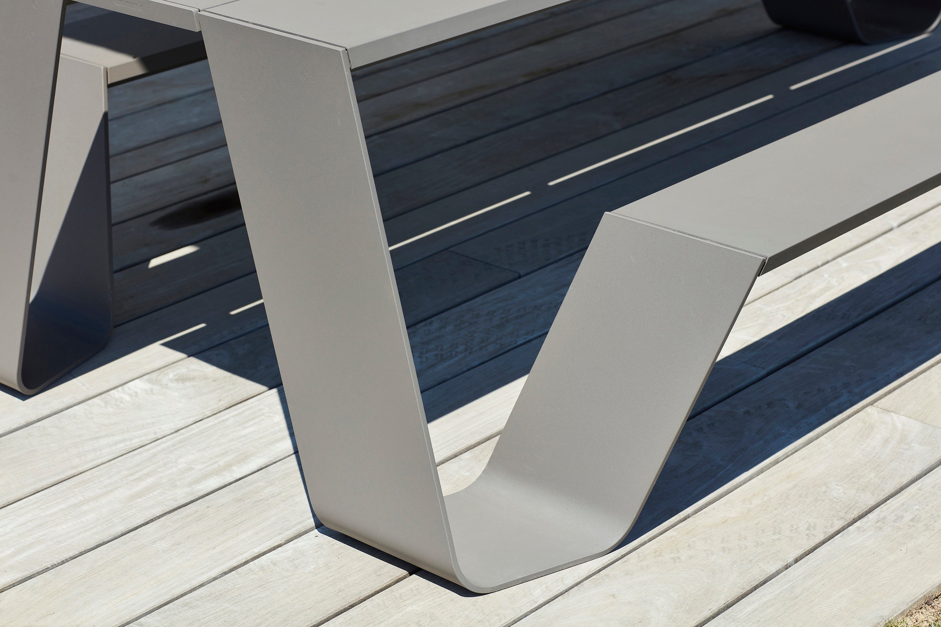 Close-up of the curved leg and bench support of a grey Hopper AA table on pale wooden decking.