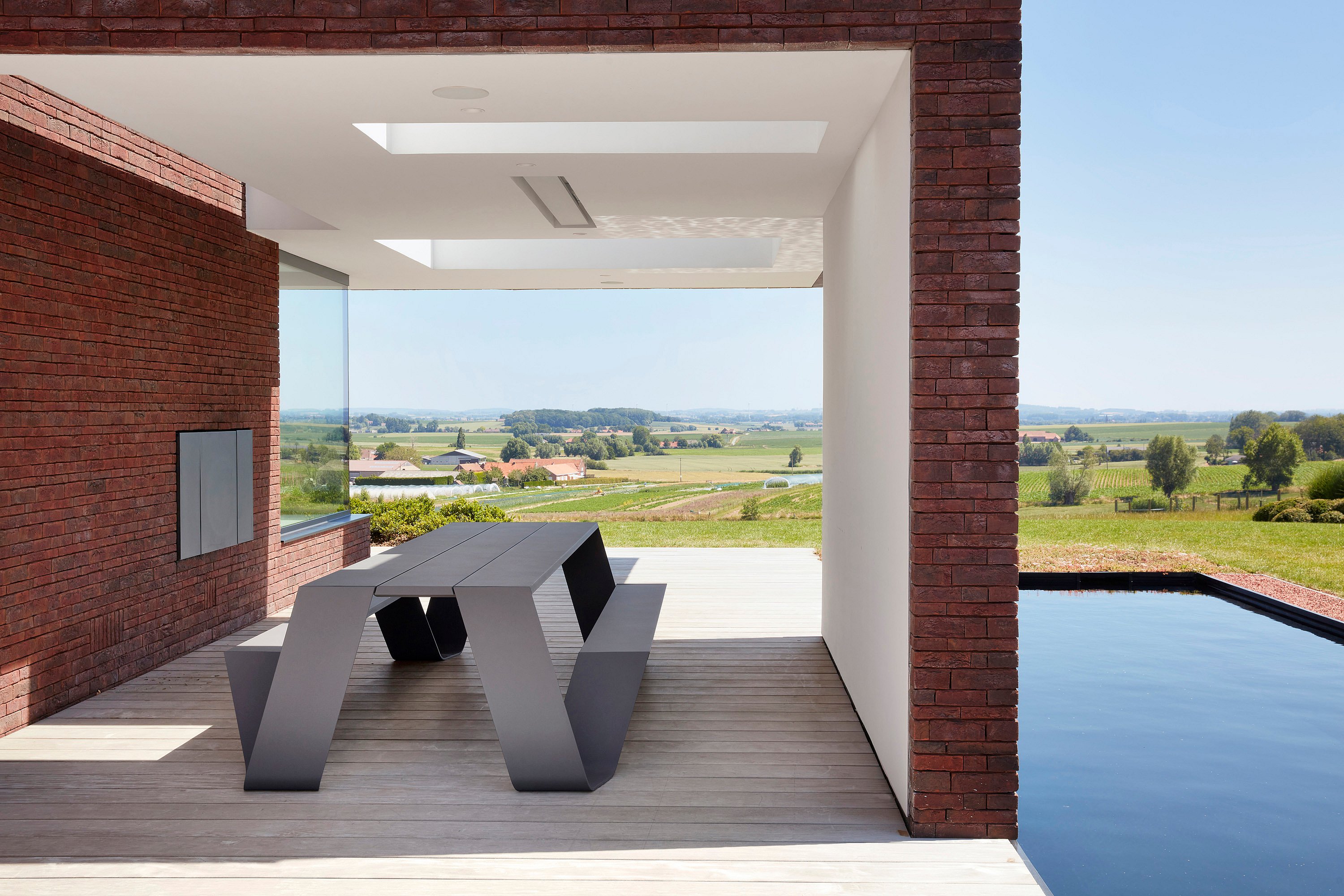 An empty grey Hopper AA table stands under a covered terrace beside a reflecting pool, framed by brick walls and a wide countryside view.