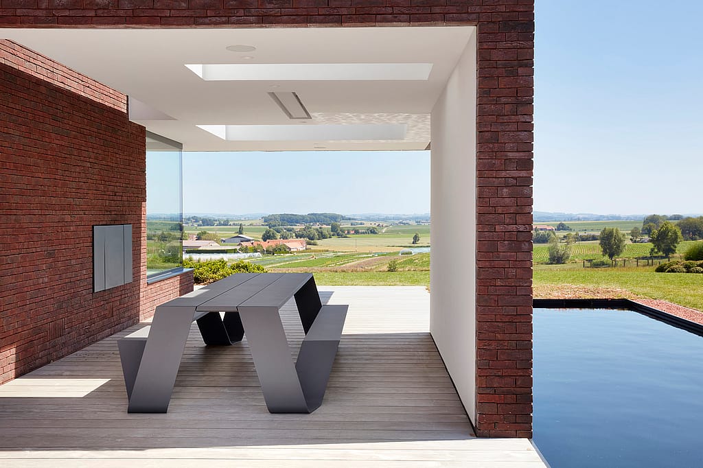 An empty grey Hopper AA table stands under a covered terrace beside a reflecting pool, framed by brick walls and a wide countryside view.
