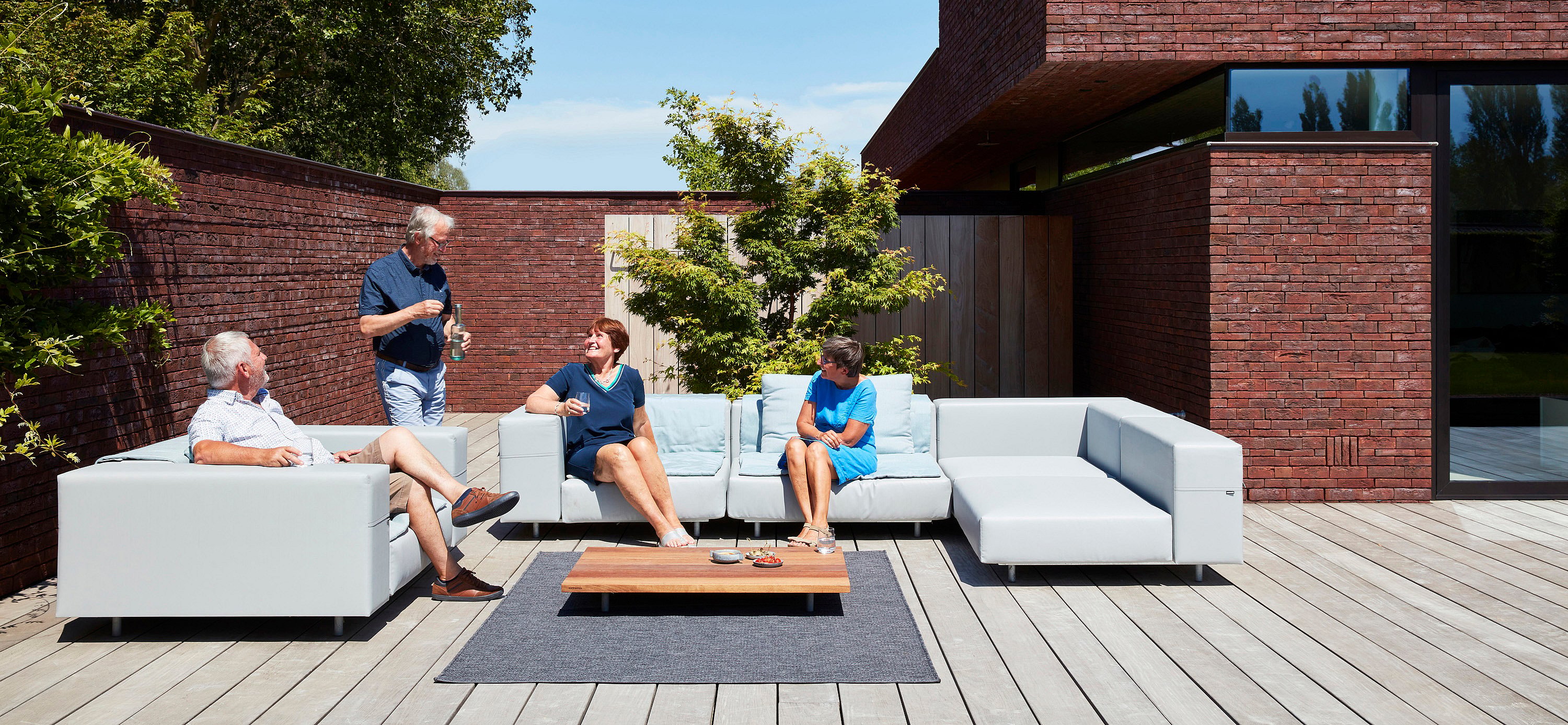 Four friends relax and chat on a light grey Walrus sofa set around a low wooden coffee table on a sunny terrace.