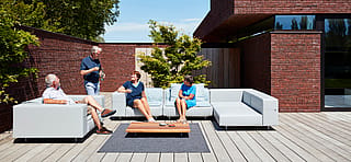 Four friends relax and chat on a light grey Walrus sofa set around a low wooden coffee table on a sunny terrace.