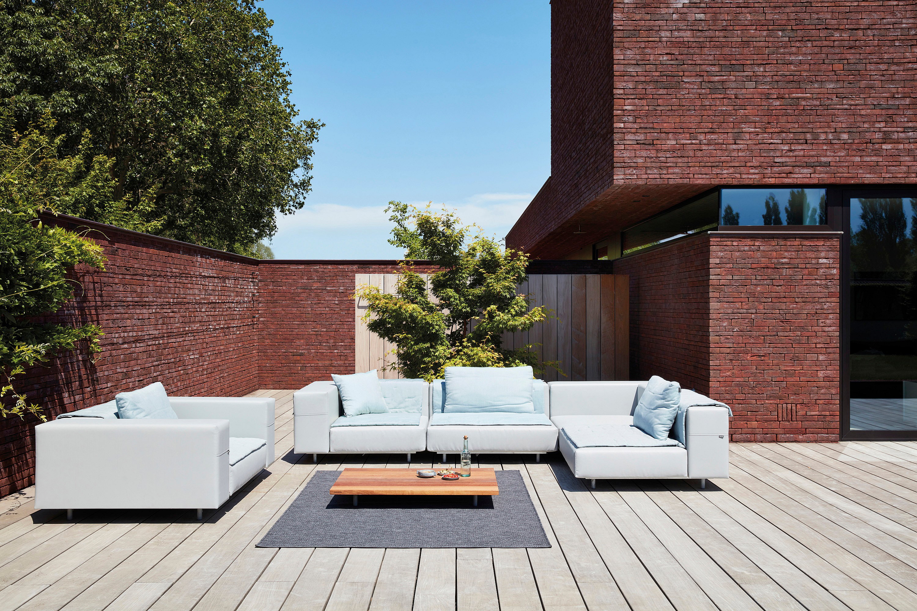 Un canapé Walrus gris clair aux coussins bleu pâle est installé sur une terrasse en bois ensoleillée, à côté d'une maison moderne en briques rouges. Une table basse en bois est posée sur un tapis d'extérieur devant.