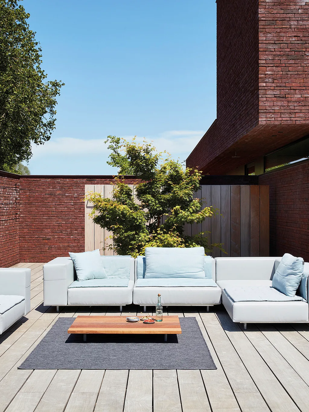 A light grey Walrus sofa set with pale blue cushions sits on a sunlit wooden terrace beside a modern red-brick house, with a low wooden coffee table on an outdoor rug in front.