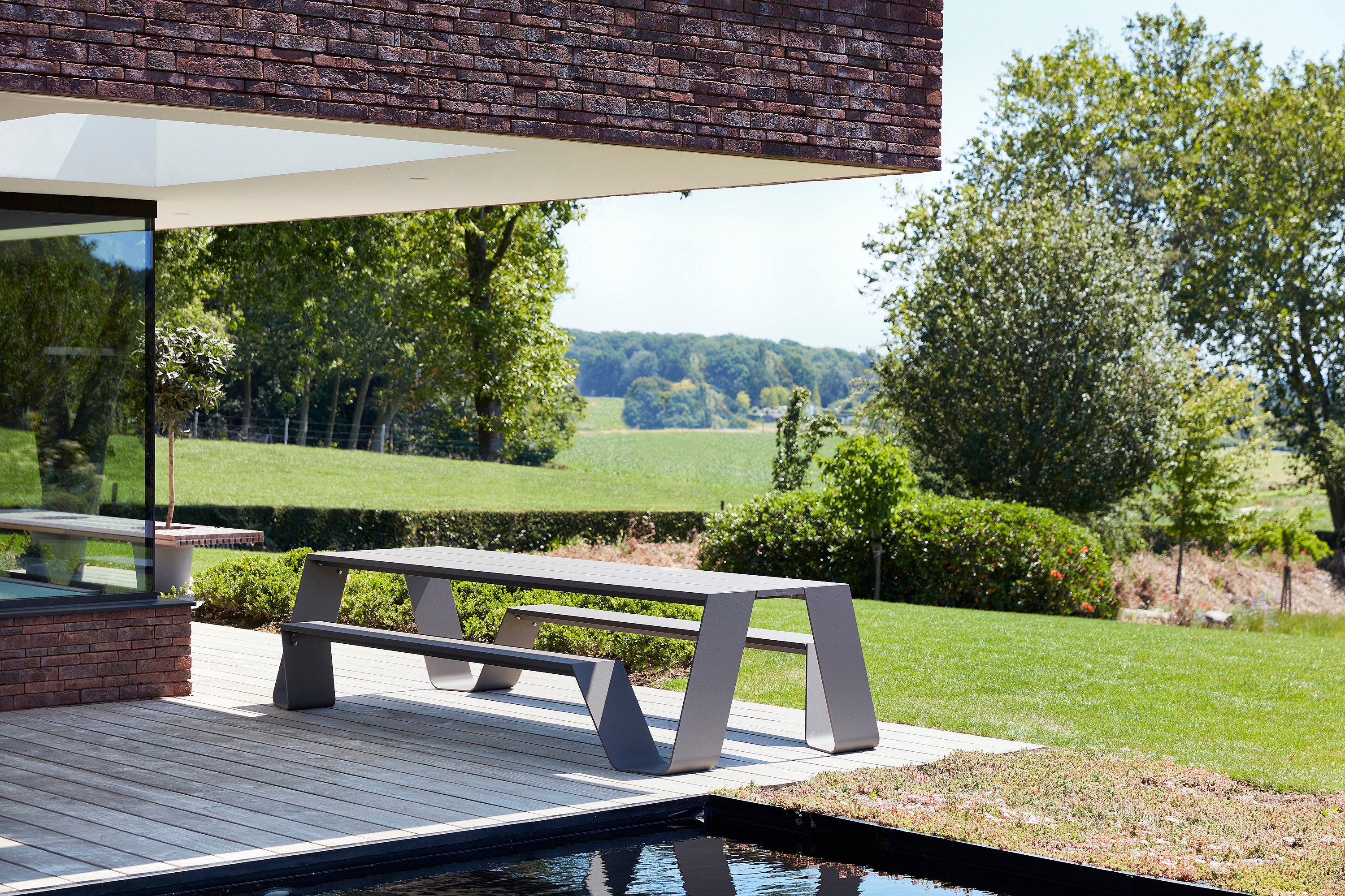 An empty grey Hopper AA table stands on the terrace beside a dark pool, overlooking a manicured lawn and open countryside.