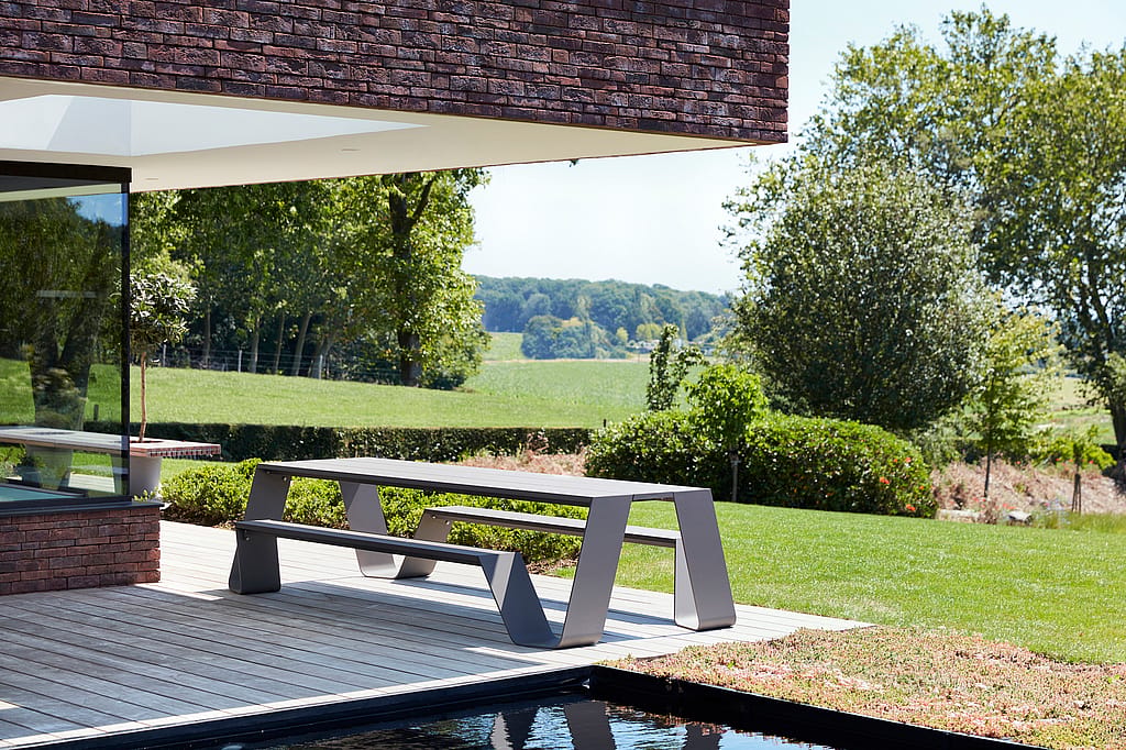 An empty grey Hopper AA table stands on the terrace beside a dark pool, overlooking a manicured lawn and open countryside.