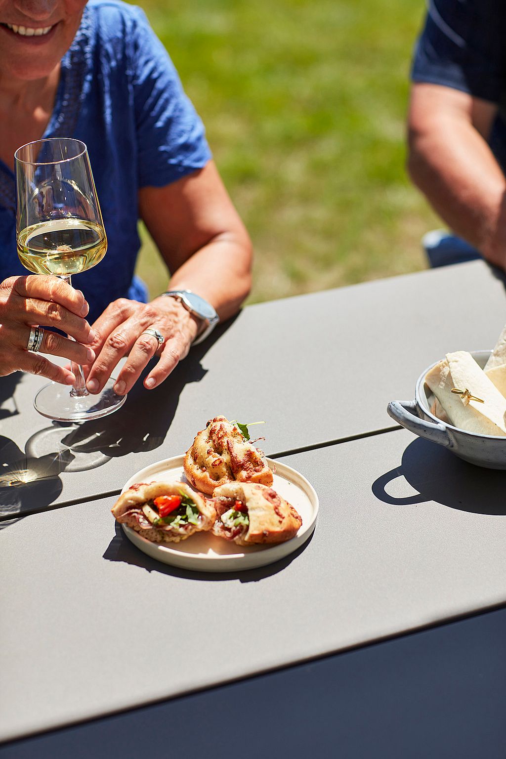 Close-up of a glass of white wine and small sandwiches on a grey Hopper AA tabletop during an outdoor lunch.