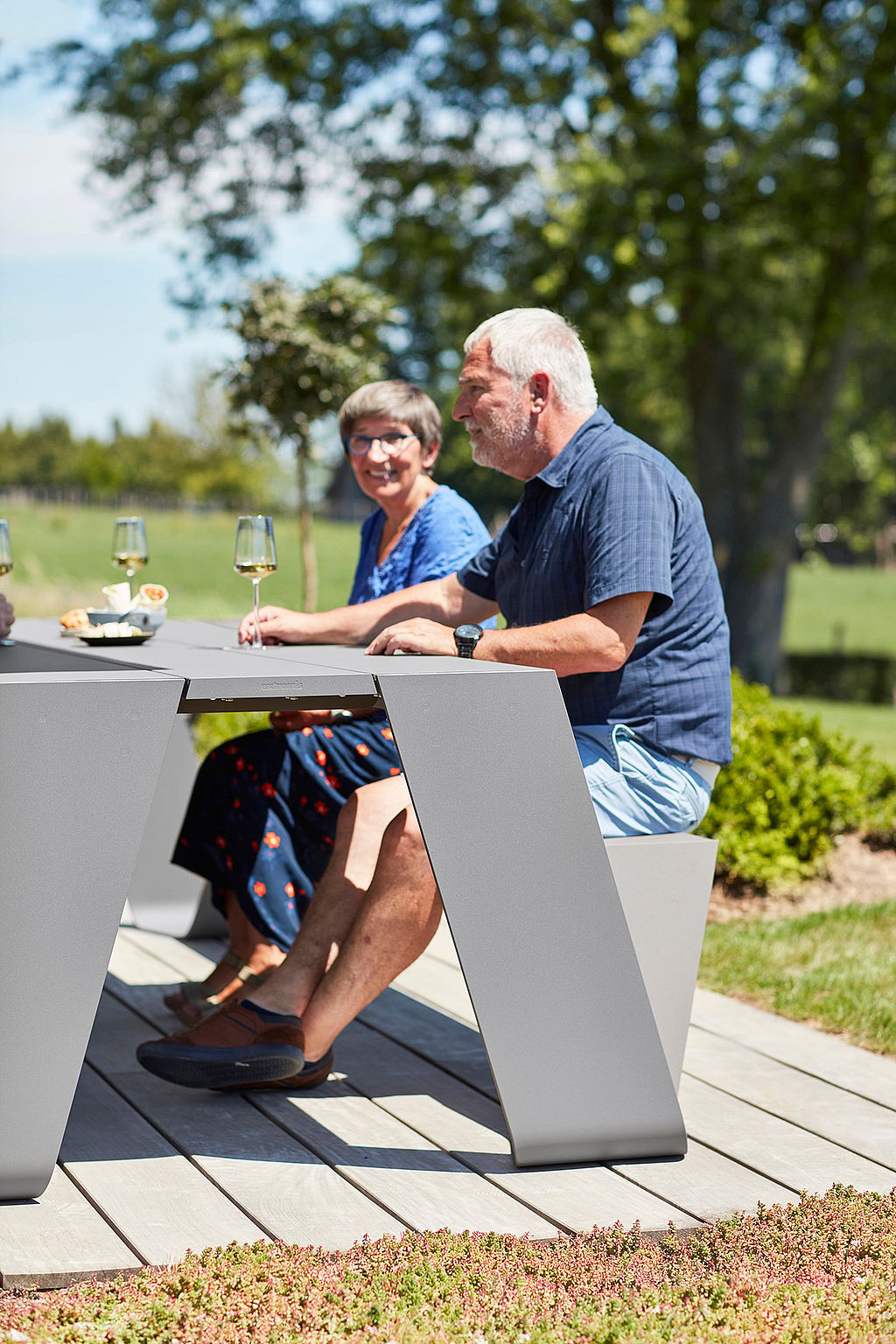 Two friends sit at a grey Hopper AA table on a sunny terrace, enjoying glasses of white wine with trees and lawn behind them.
