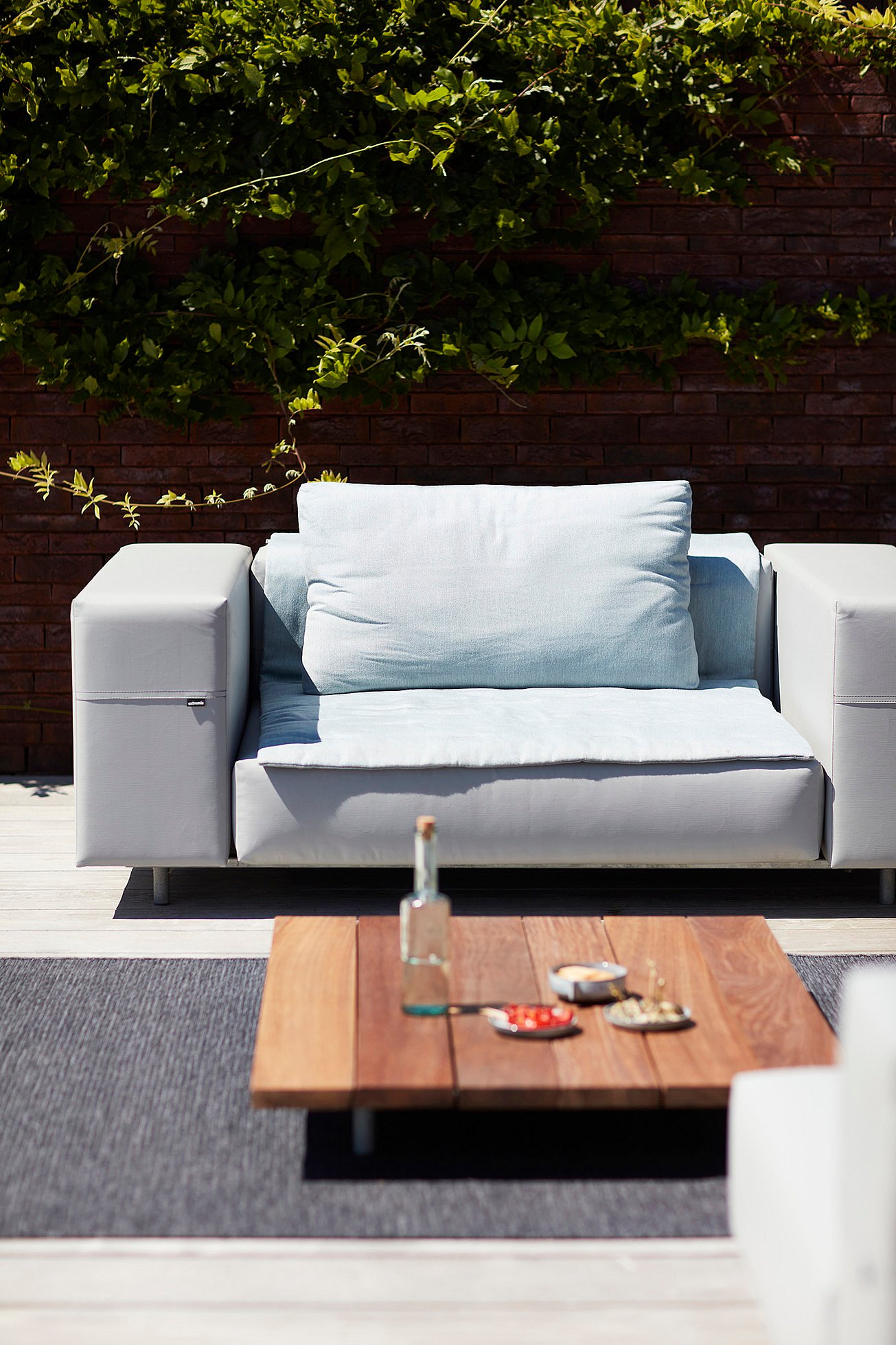 Close-up of a single light grey Walrus seat with a pale blue cushion, set behind a low wooden coffee table against a brick wall covered with greenery.