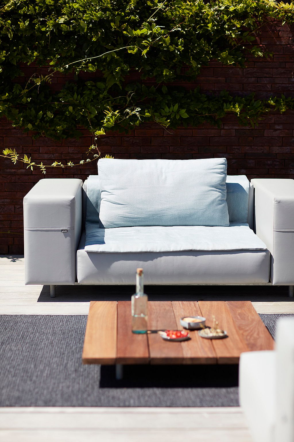 Close-up of a single light grey Walrus seat with a pale blue cushion, set behind a low wooden coffee table against a brick wall covered with greenery.