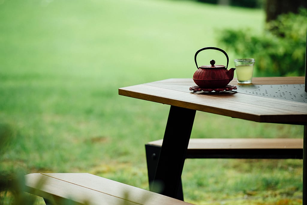 Close-up of a red teapot and a glass of green tea on the corner of a wooden outdoor anker table, with a soft-focus lawn in the background.