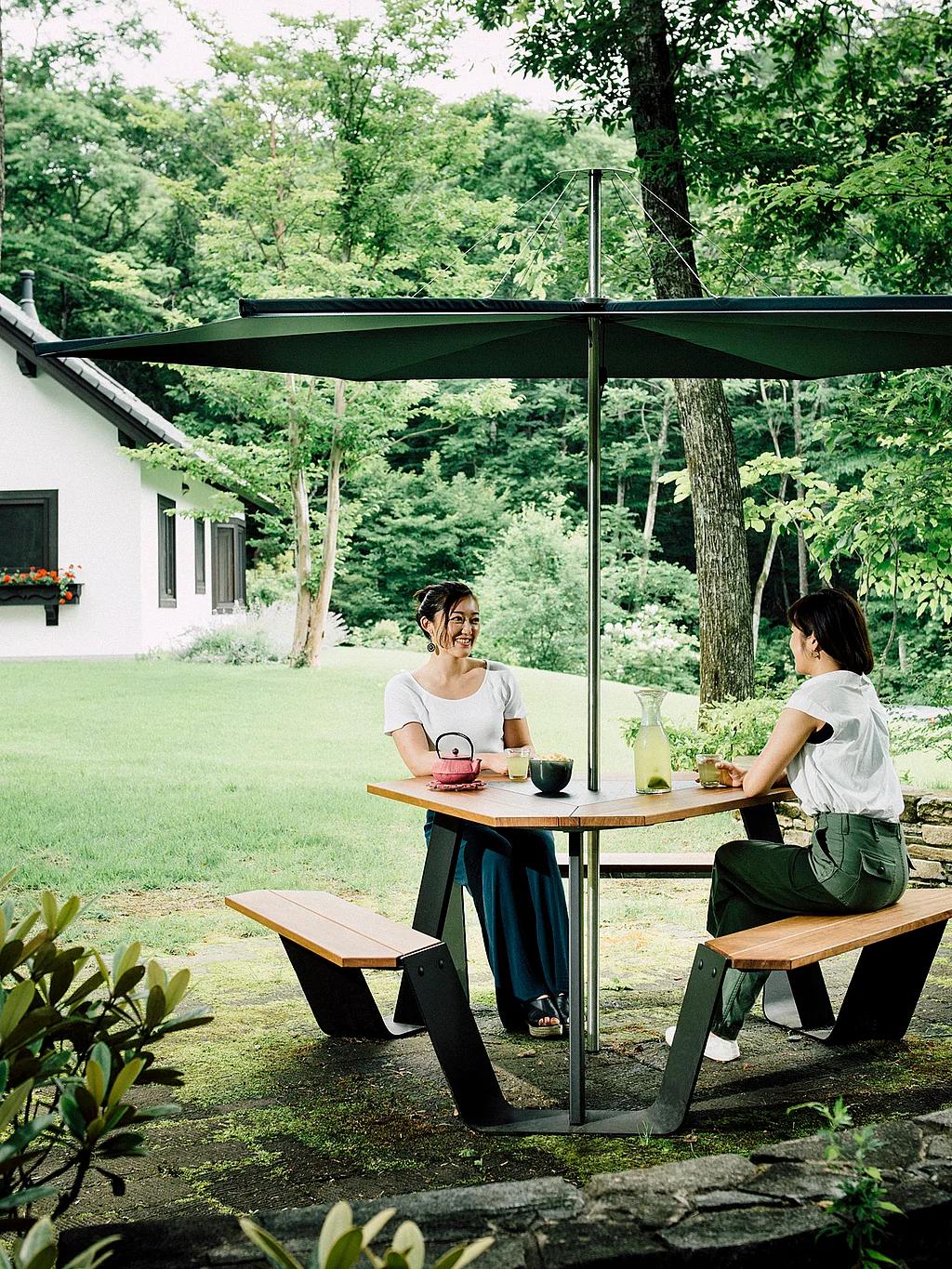 Zwei Frauen trinken Tee an einem hölzernen Ankertisch mit angebrachten Bänken unter einem großen dunklen Inumbrina-Sonnenschirm in einem üppigen Garten neben einem weißen Haus.