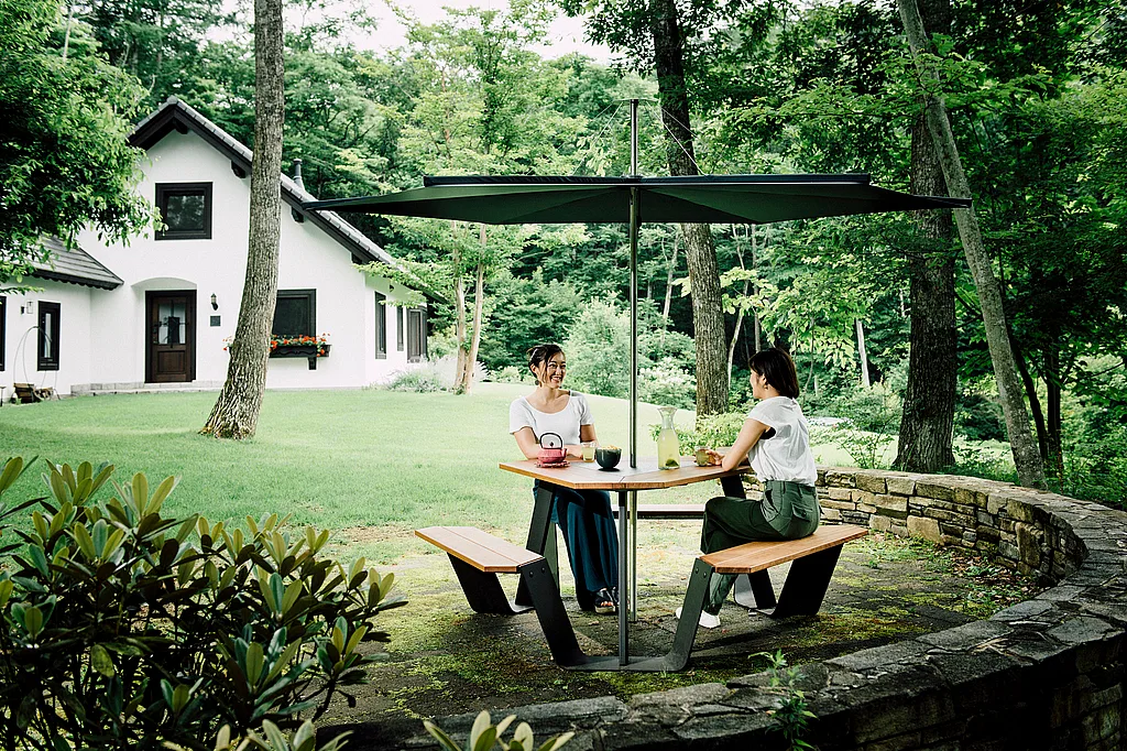 Two women having tea at a wooden anker table with attached benches under a large dark inumbrina parasol in a lush garden beside a white house.