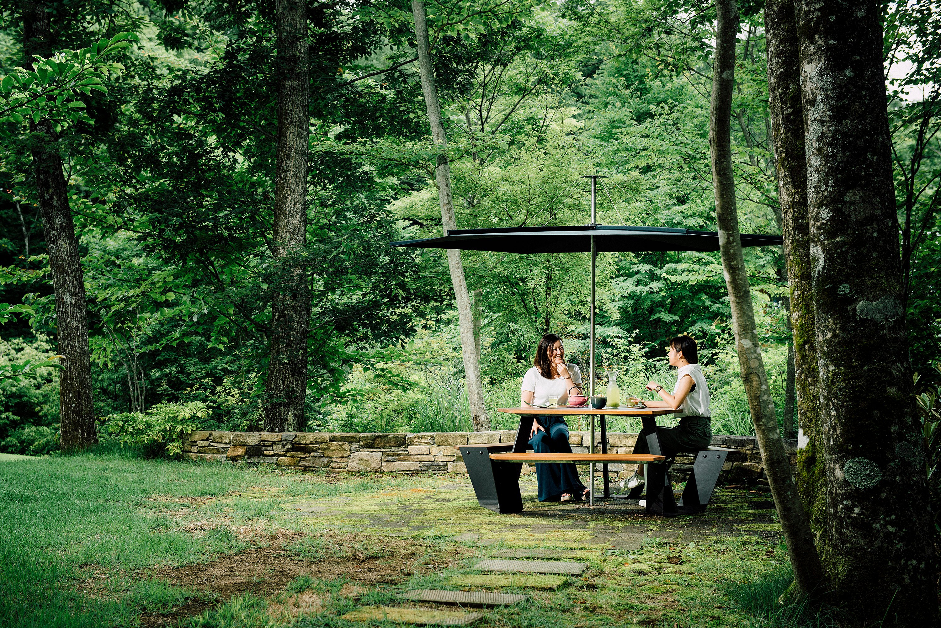 Twee mensen aan een zwarte Anker-picknicktafel met overkapping in een bosrijke tuin.