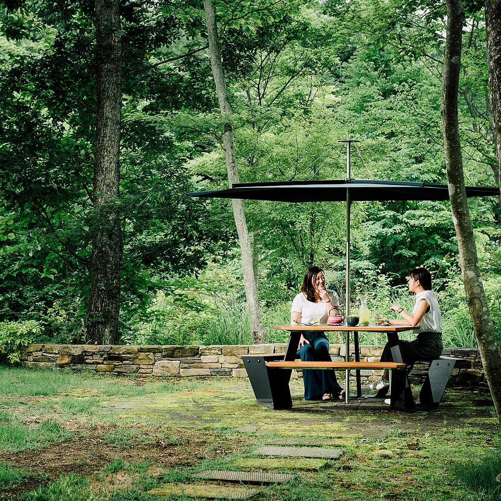 Two people sitting at a black Anker picnic table with canopy in a wooded garden.