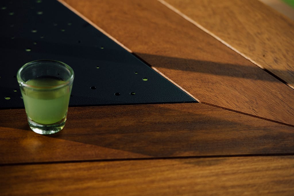 Small glass of green tea resting on a geometric wooden tabletop with a black perforated metal center panel and angled sunlight across the surface.