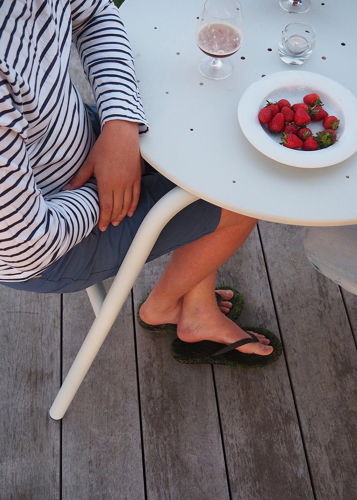 Close-up of a light blue round Virus table with a plate of strawberries, glasses and part of a seated person in a striped shirt, seen against weathered wooden decking.
