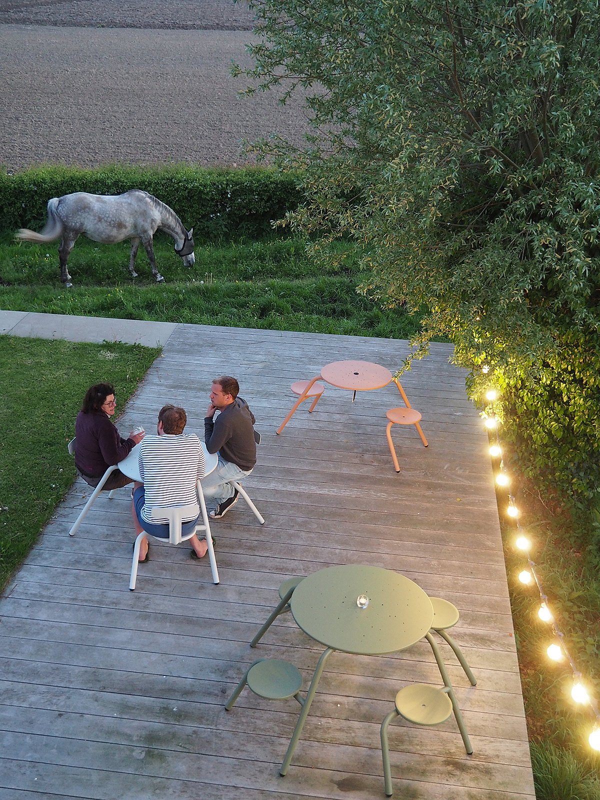 Overhead view of a large wooden platform in the countryside with three Virus picnic tables in different colors, three people talking at one table, a horse grazing nearby and string lights along the edge.