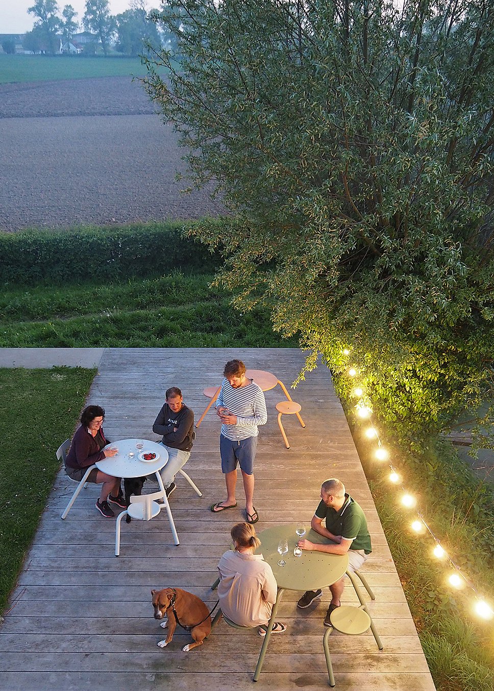 Abendansicht von oben auf eine hölzerne Gartenterrasse mit mehreren runden Virus-Picknicktischen in verschiedenen Farben, an denen sich Menschen mit Getränken neben einem Hund und einer Reihe leuchtender Lichterketten versammeln.