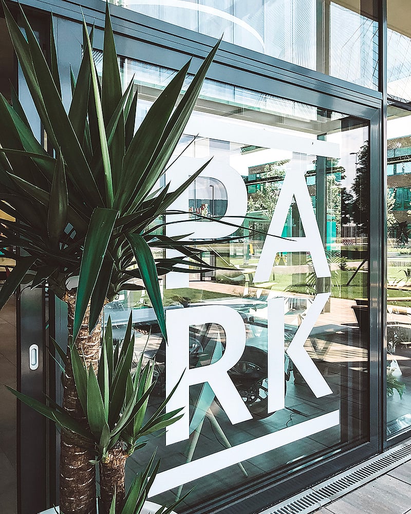   Große weiße Buchstaben „PARK“ prangen auf der Glasfassade. Im Vordergrund steht eine hohe, stachelige Pflanze, und die Terrasse spiegelt sich im Fenster.