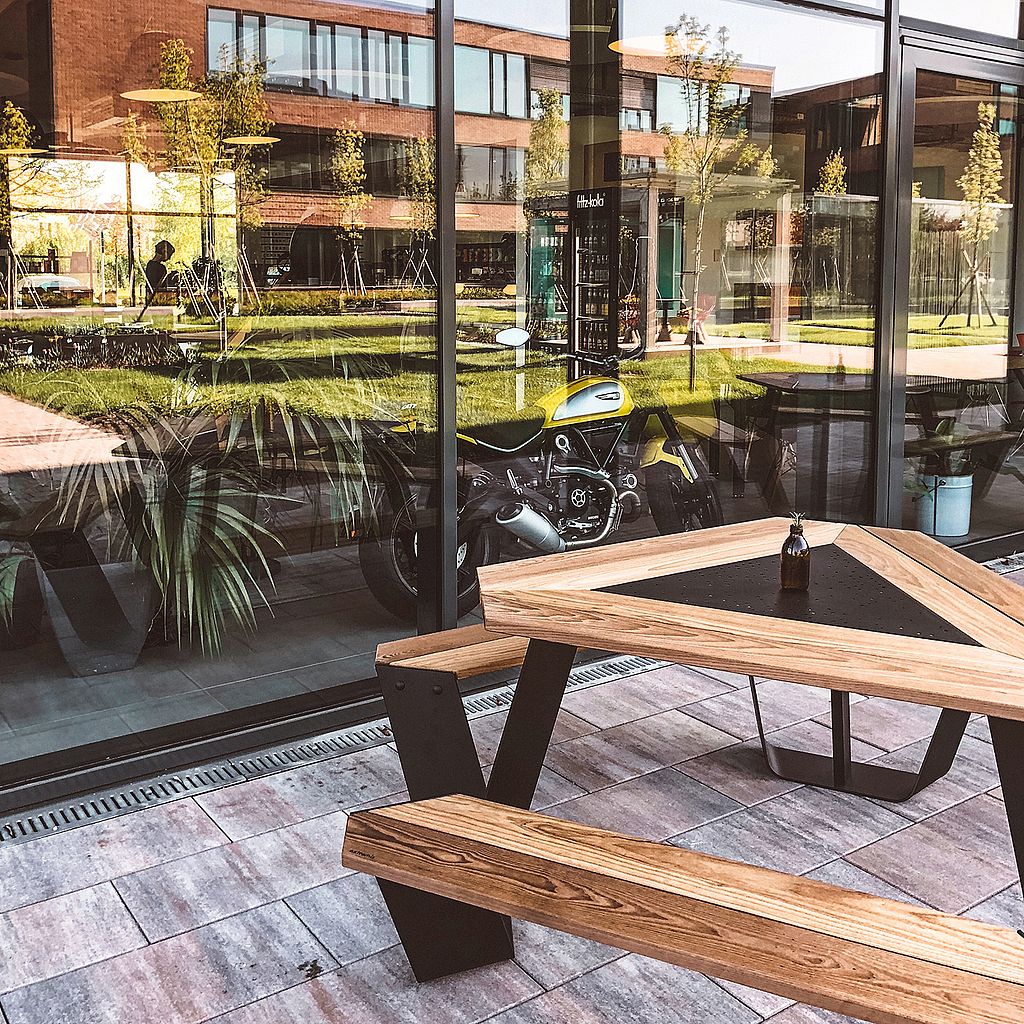 Ein Anker-Tisch mit Holzplatte und integrierten Bänken steht neben dem Fenster des Cafés. Im Inneren spiegeln sich die begrünte Büroanlage und ein gelbes Motorrad.