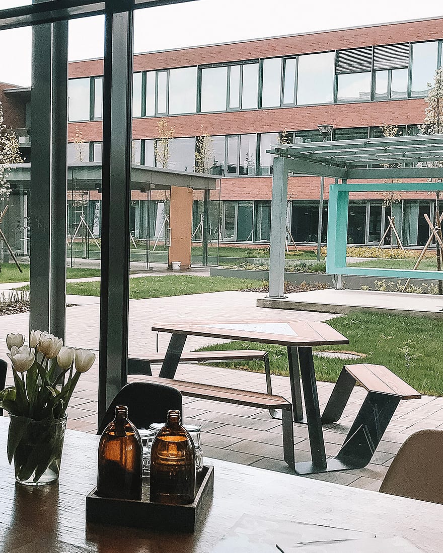 Blick aus dem Café auf einen einzelnen Anker-Tisch im Freien. Tulpen, bernsteinfarbene Flaschen und der Bürokomplex sind durch die großen Fenster zu sehen.
