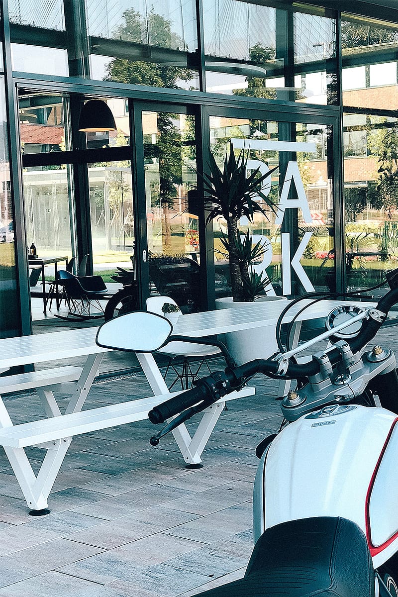 Ein weißer Marina-Picknicktisch steht vor der Glasfassade des Park Coffee & Kitchen. Im Vordergrund sind das große PARK-Logo im Schaufenster und ein Motorrad zu sehen.