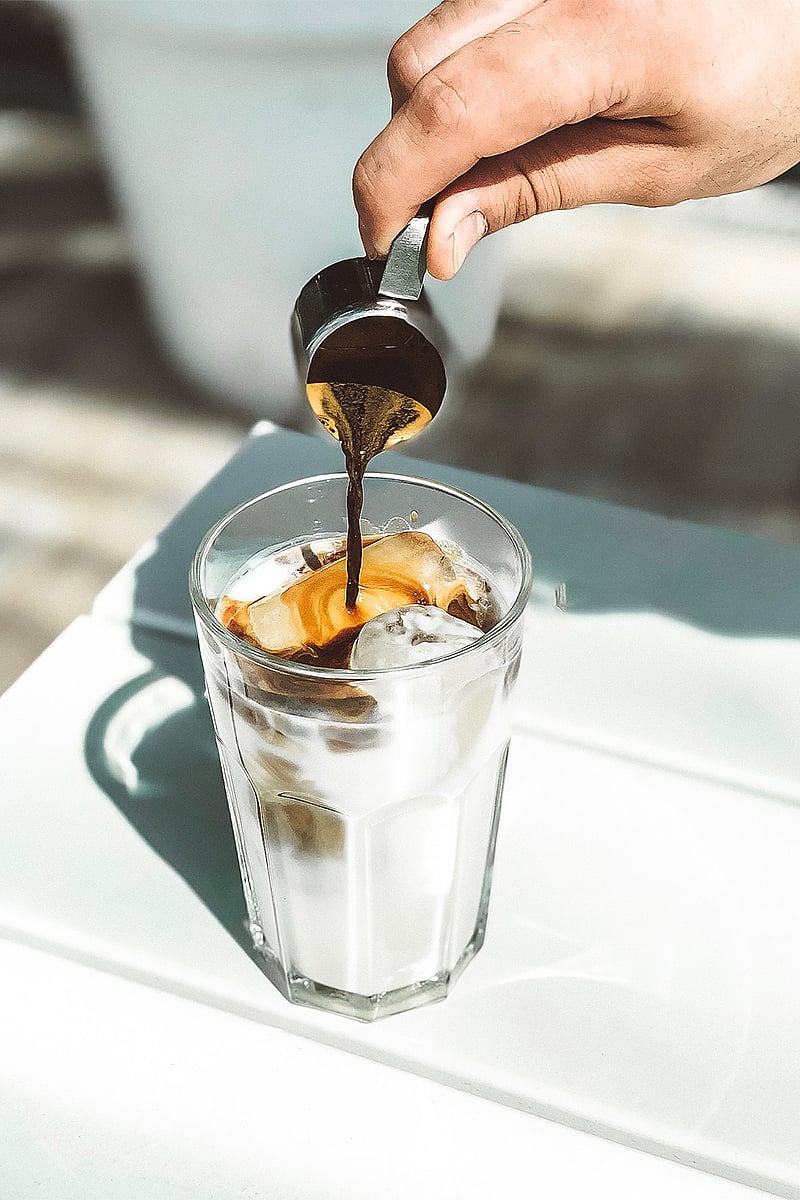  Nahaufnahme: Espresso wird in hellem Sonnenlicht über Eis in ein Glas Milch auf einem weißen Tisch gegossen.