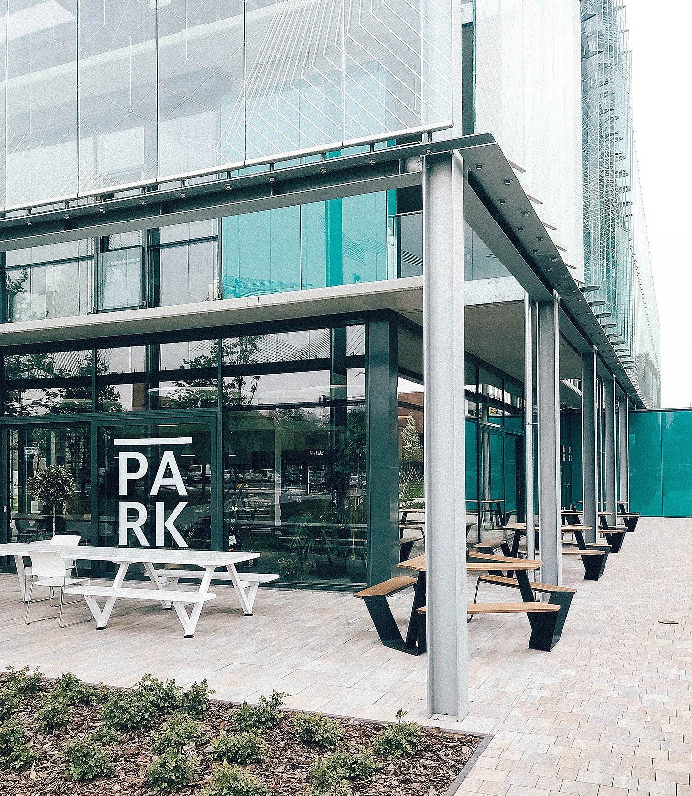 Außenansicht des Park Coffee & Kitchen, eines Cafés mit Glasfassade im Graphisoft Park, mit einem weißen Marina-Picknicktisch und mehreren Anker-Tischen mit Holzplatte auf der überdachten Terrasse.