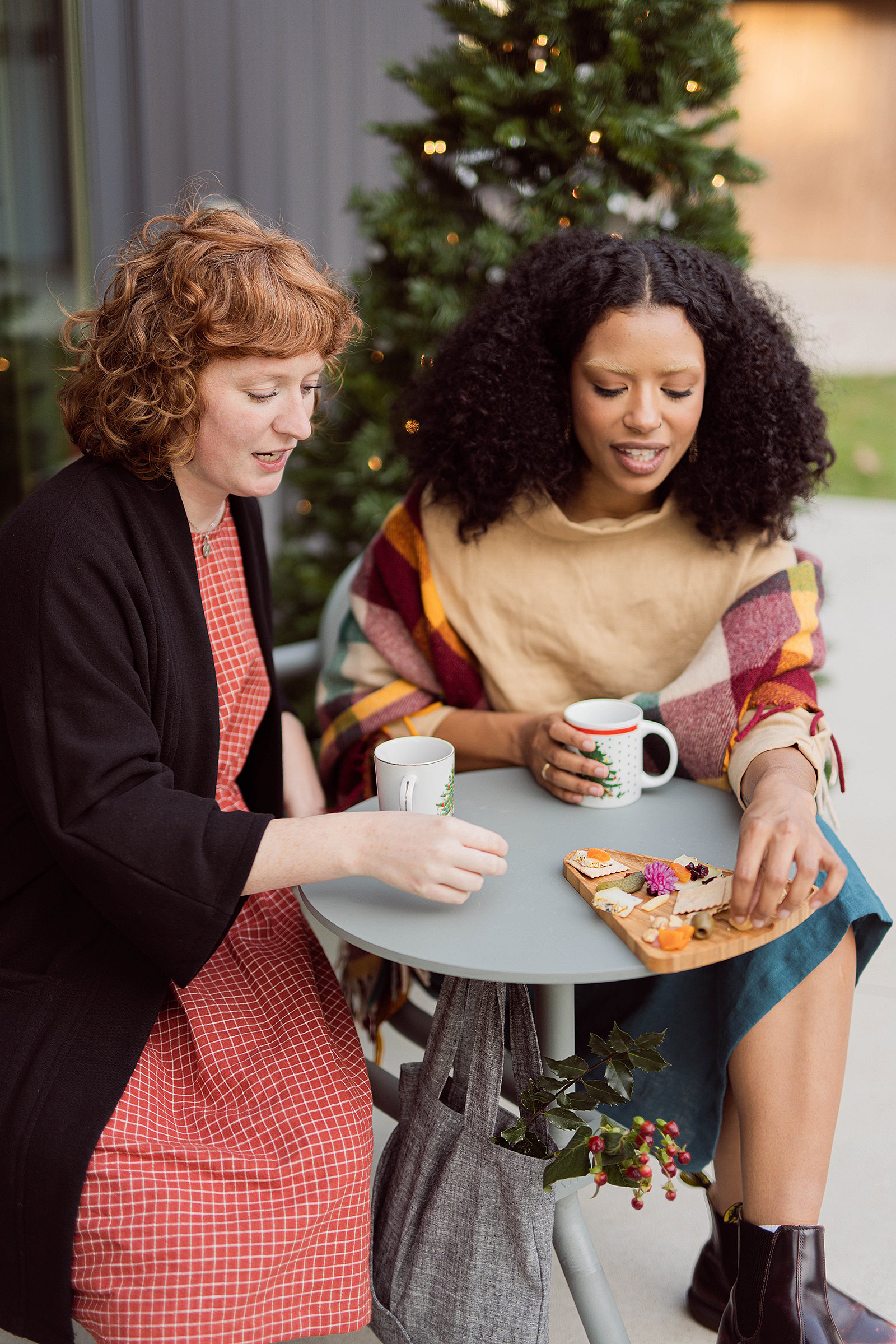 Close-up van twee vrouwen die snacks en warme dranken delen aan een ronde Bistroo-tafel, met een draagtas eronder en een verlichte boom op de achtergrond.