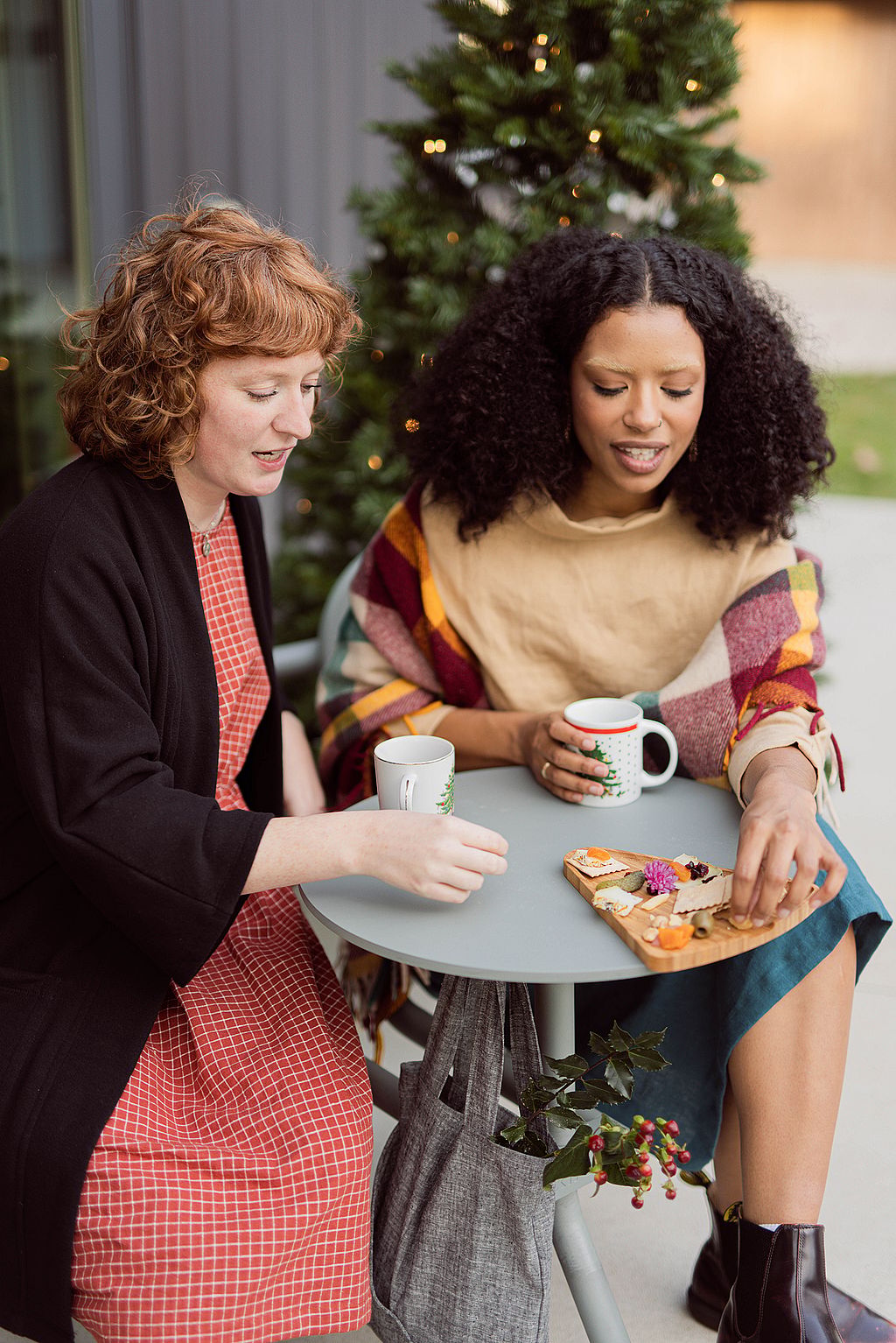 Close-up van twee vrouwen die snacks en warme dranken delen aan een ronde Bistroo-tafel, met een draagtas eronder en een verlichte boom op de achtergrond.
