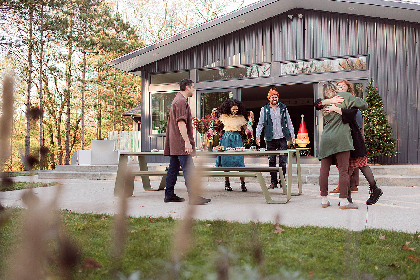 Een groep vrienden verzamelt zich rond een groene Picnik-tafel buiten een in het donker gekleed modern huis, met een paar knuffels en anderen die eten en drinken bereiden.