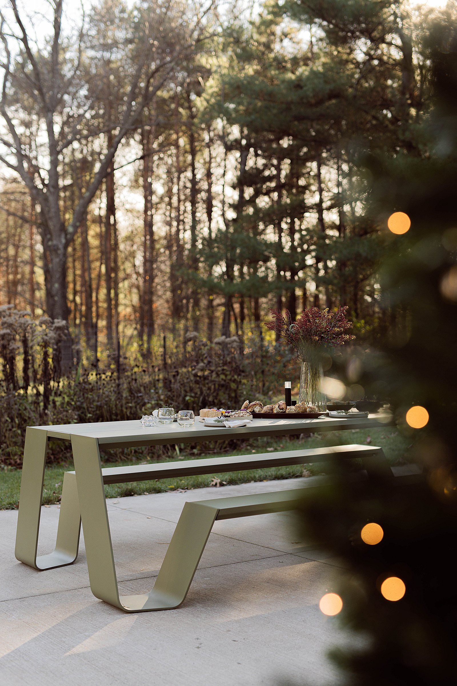 Groene Picnik-tafelset voor een buitenmaaltijd op een terras in de schemering, met bomen achter en warme wazige lichten van een nabijgelegen versierde boom op de voorgrond.