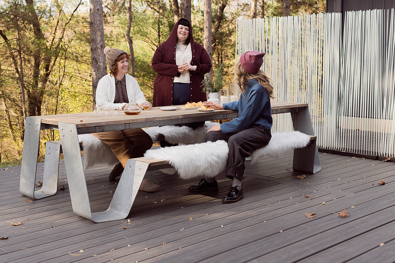 Drie vrouwen kletsen rond een houten Hopper-picknicktafel met witte schapenvachtbankhoezen, gebak en thee, op een bosdek.