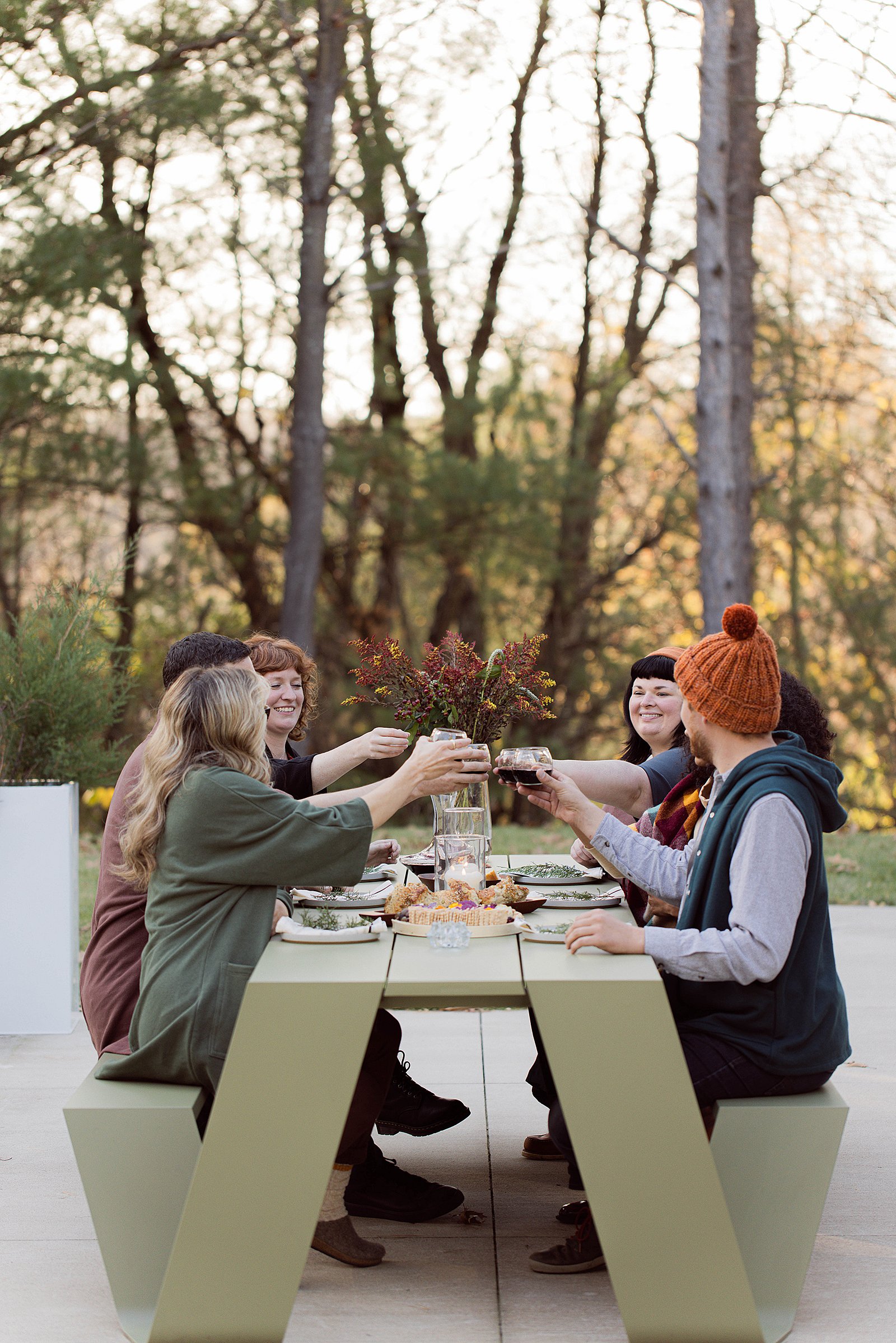 Drie vrouwen verzamelen zich rond een houten hoppenpicknicktafel op een dek naast een modern huis in het bos, met gebak en thee op tafel en een edelhertsculptuur op het gazon.
