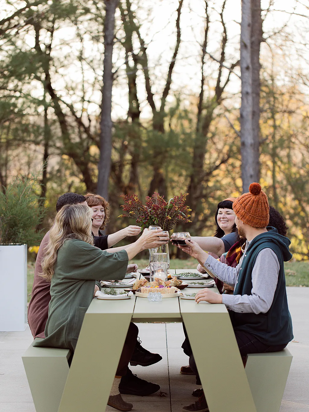 Drie vrouwen verzamelen zich rond een houten hoppenpicknicktafel op een dek naast een modern huis in het bos, met gebak en thee op tafel en een edelhertsculptuur op het gazon.