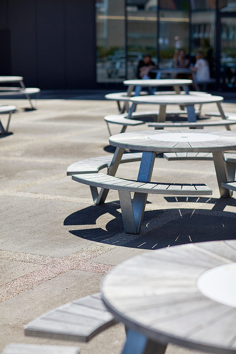 Gros plan d'une rangée de tables de pique-nique Pantagruel vides sur la terrasse ensoleillée, devant la façade vitrée du bâtiment du campus.