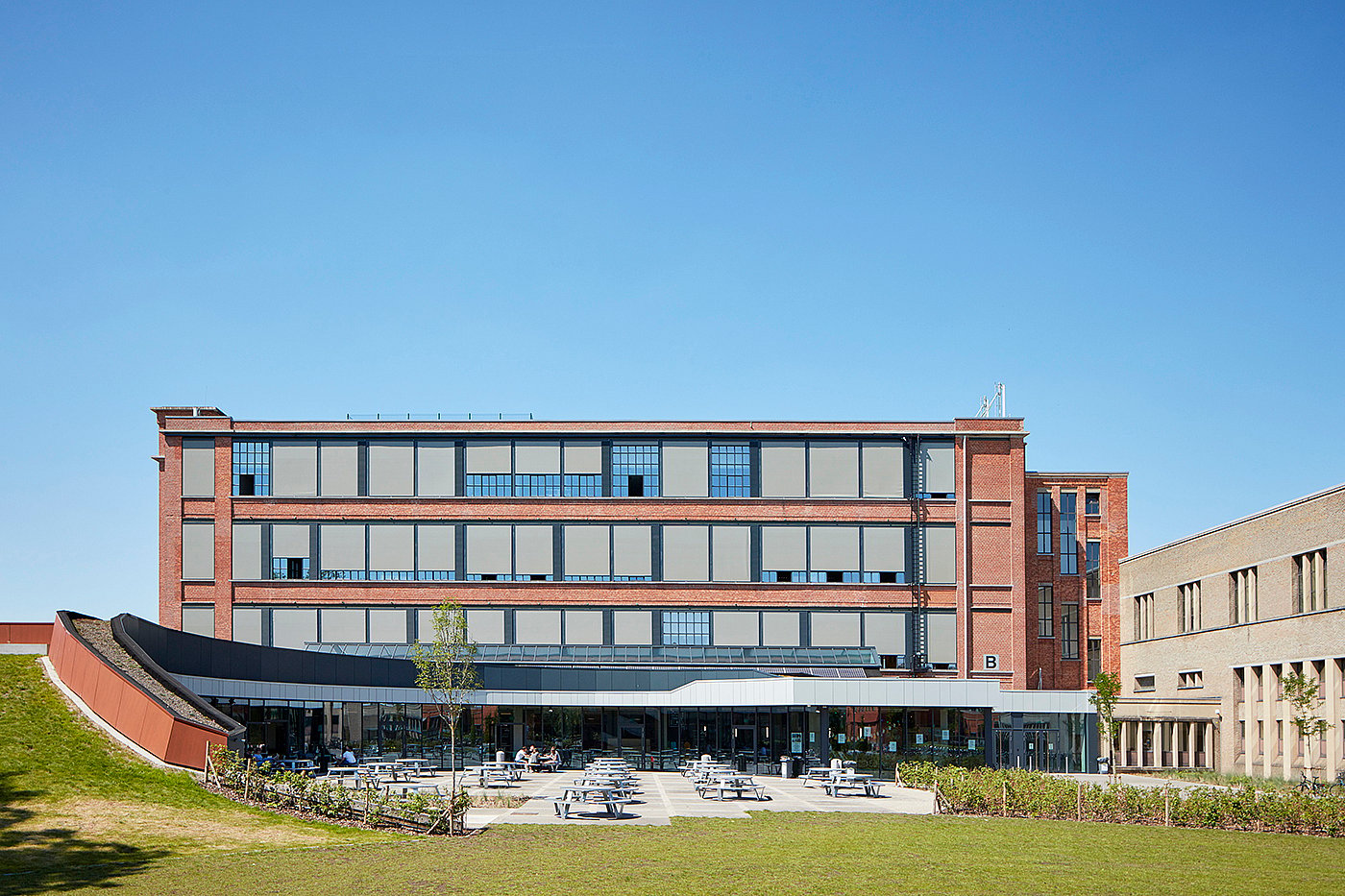   Vue extérieure large de l'Odisee University College, avec une grande terrasse équipée de tables de pique-nique Pantagruel devant le bâtiment moderne en briques et en verre.