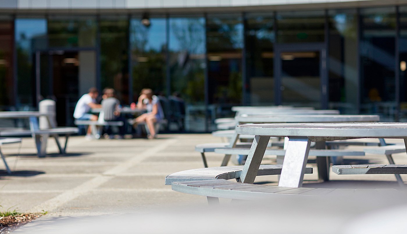 Détail en contre-plongée de plusieurs tables de pique-nique Pantagruel à l'extérieur du bâtiment du campus, avec des étudiants rassemblés à une table, flous en arrière-plan.