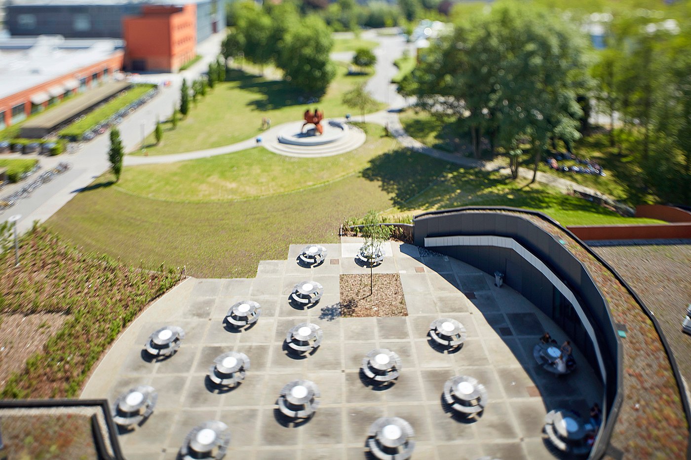 Vue aérienne en tilt-shift de la terrasse du campus d'Odisee, avec plusieurs tables de pique-nique Pantagruel, bordée de toits verts, d'une architecture aux formes courbes et d'une grande pelouse au-delà.