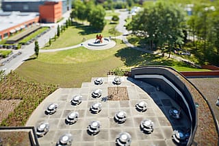 Vue aérienne en tilt-shift de la terrasse du campus d'Odisee, avec plusieurs tables de pique-nique Pantagruel, bordée de toits verts, d'une architecture aux formes courbes et d'une grande pelouse au-delà.