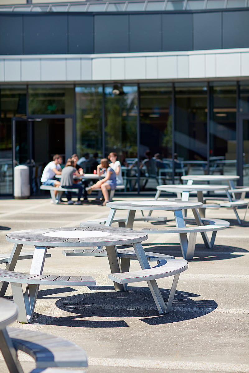 Plusieurs tables de pique-nique Pantagruel sont installées sur la terrasse extérieure d'Odisee, tandis qu'un groupe d'étudiants est assis près de l'entrée, à l'arrière-plan.