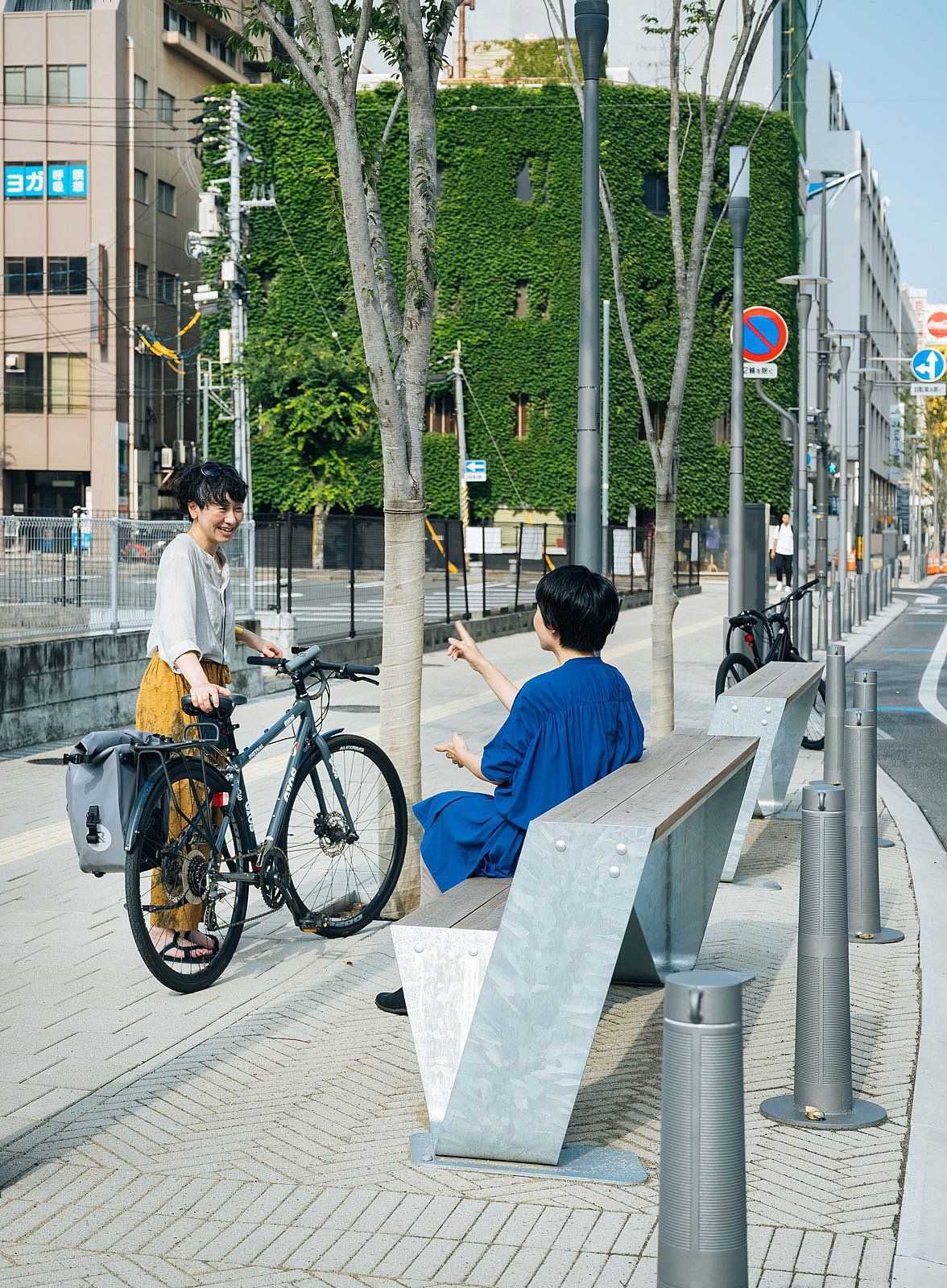 Zwei Personen unterhalten sich neben einer Hopper-Bank aus verzinktem Stahl und Holz auf einem breiten Bürgersteig der Stadt, eine Person steht neben einem Fahrrad und im Hintergrund stehen mit Efeu bedeckte Gebäude.