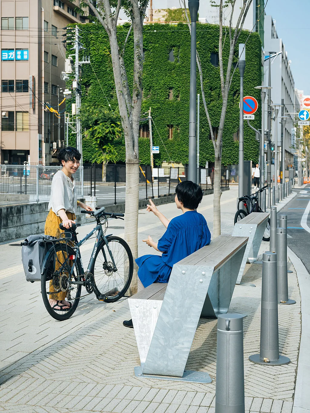 Zwei Personen unterhalten sich neben einer Hopper-Bank aus verzinktem Stahl und Holz auf einem breiten Bürgersteig der Stadt, eine Person steht neben einem Fahrrad und im Hintergrund stehen mit Efeu bedeckte Gebäude.