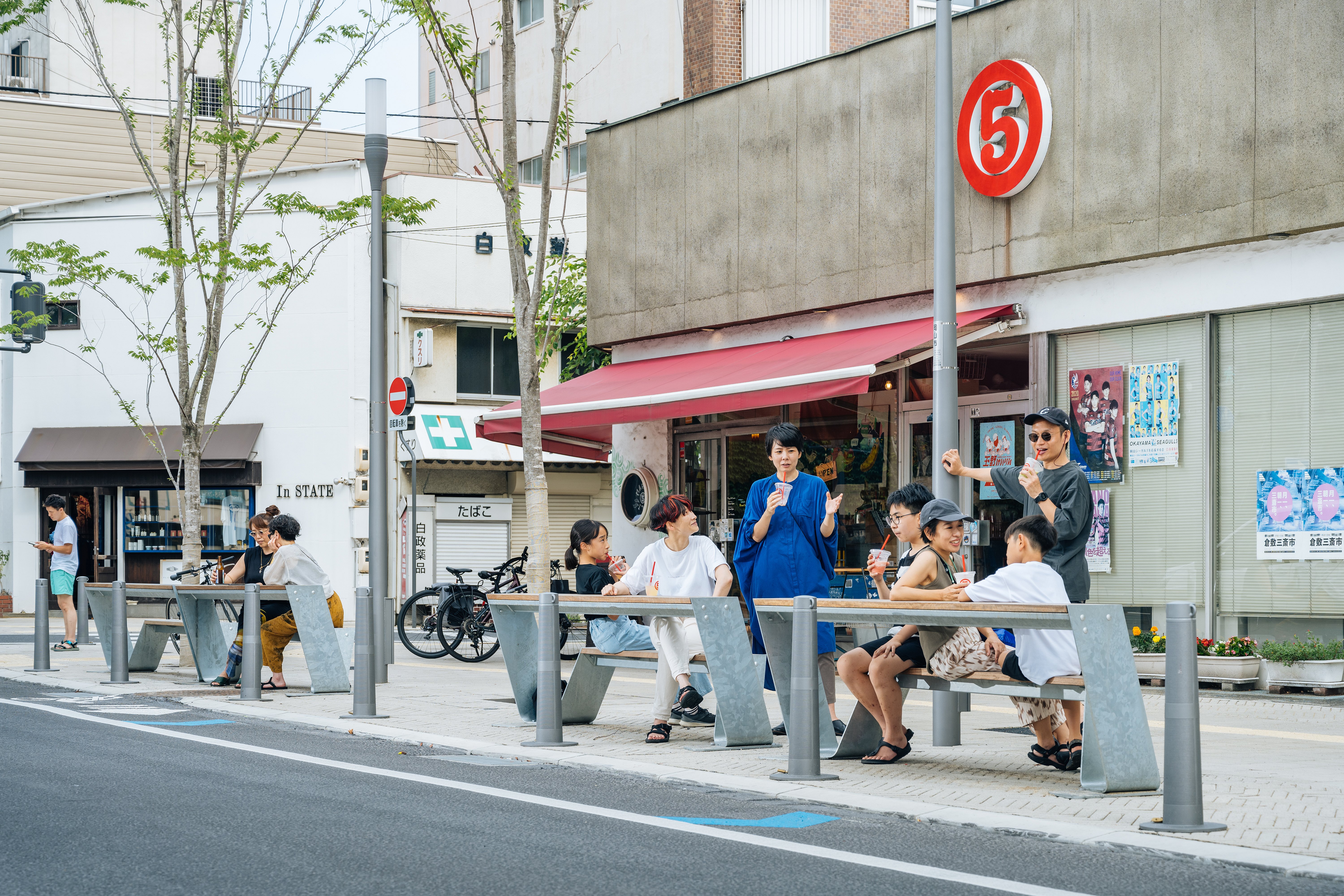 Gruppen von Menschen sitzen und unterhalten sich auf einer Reihe von Hopper-Bänken entlang einer Stadtstraße vor Geschäften und Cafés in Okayama.
