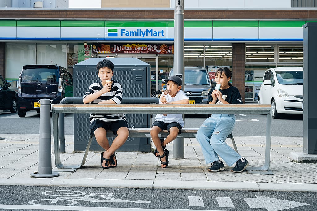 Drei Kinder essen Eis, während sie an einem langen Hopper-Banktisch vor einem FamilyMart-Supermarkt sitzen