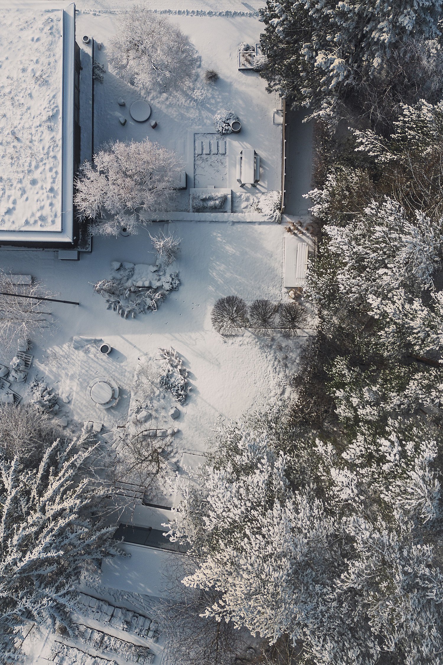 Luftaufnahme eines schneebedeckten Gartens neben einem modernen Haus mit Wegen, Pflanzbeeten und mehreren Sitzbereichen im Freien, eingebettet zwischen Bäumen.