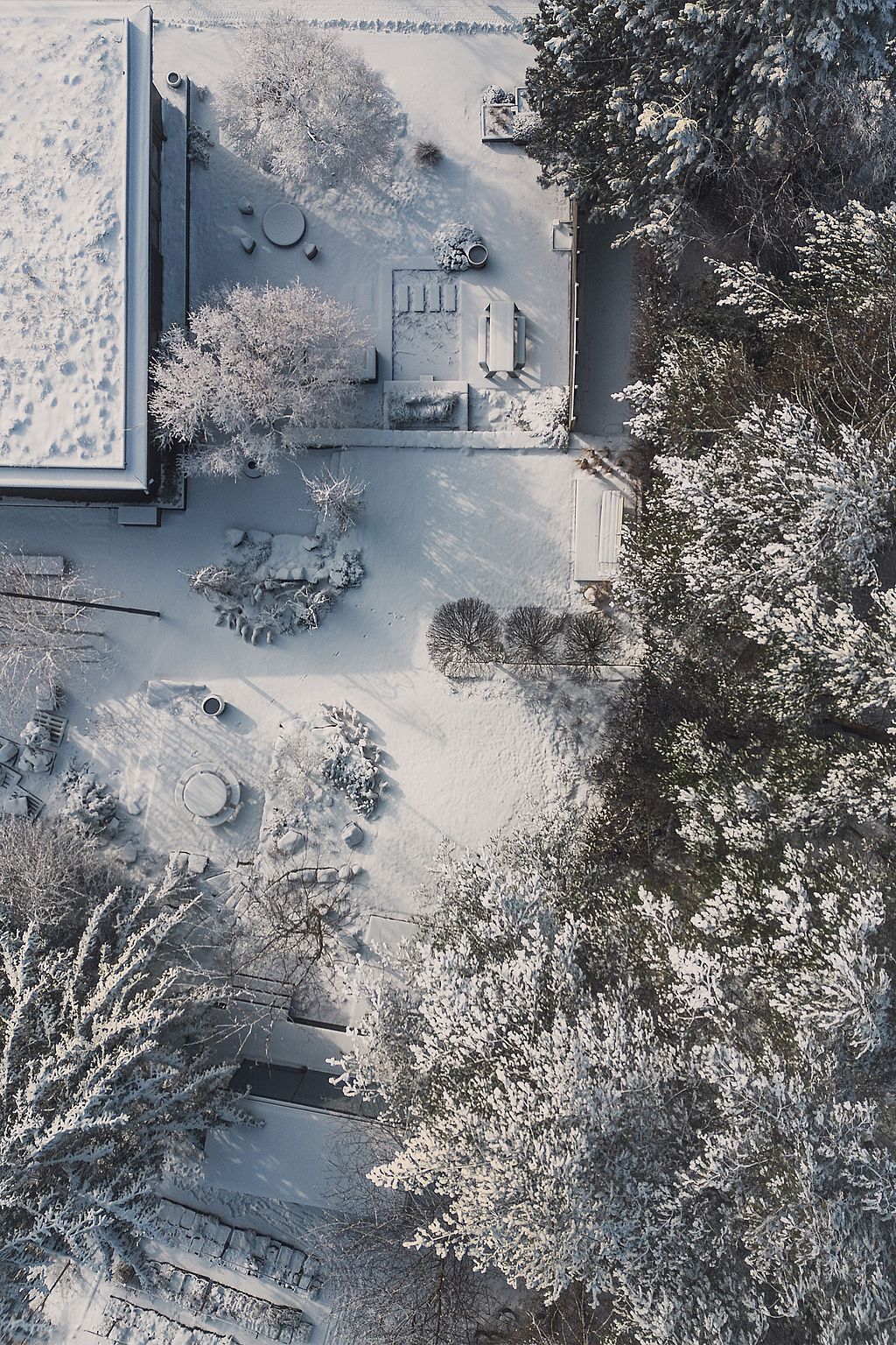 Luftaufnahme eines schneebedeckten Gartens neben einem modernen Haus mit Wegen, Pflanzbeeten und mehreren Sitzbereichen im Freien, eingebettet zwischen Bäumen.