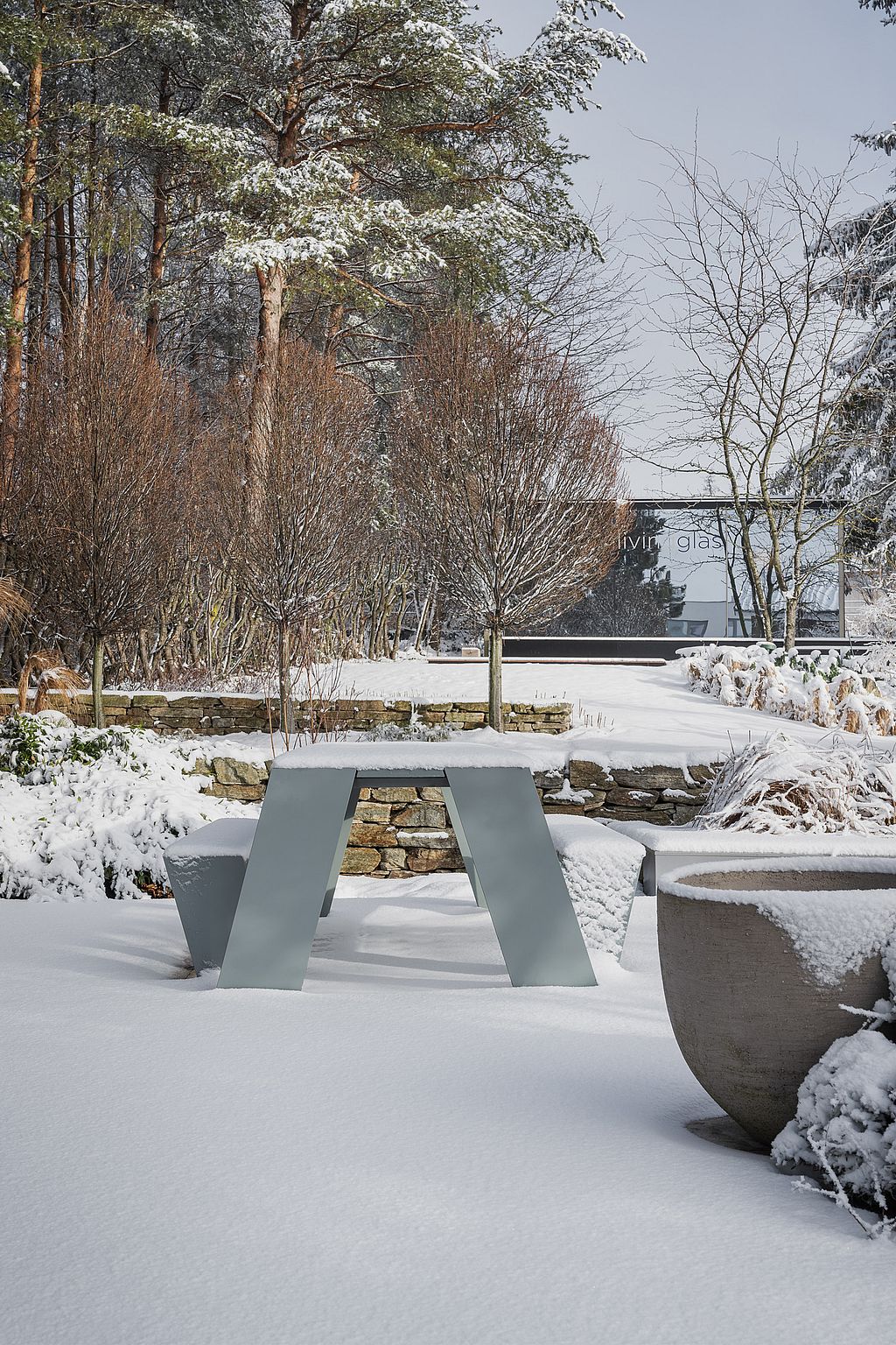 Minimaler Trichtertisch im Freien, der im tiefen Schnee neben einer Steinmauer steht, mit kahlen Wintersträuchern, hohen Kiefern und einem Gartenausstellungsgebäude im Hintergrund.