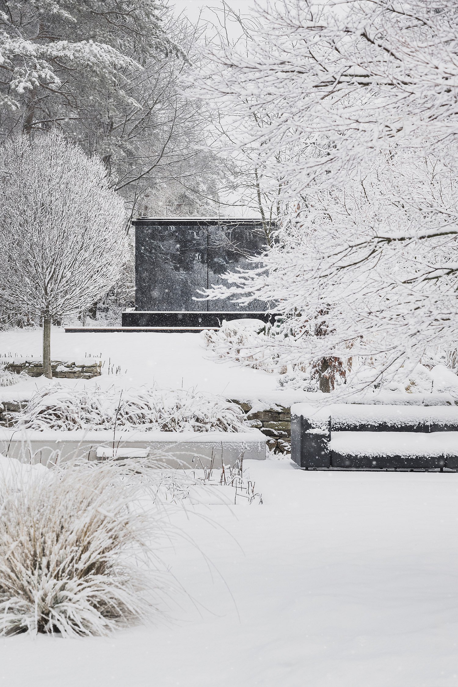 Verschneiter Wintergarten mit dunklem Außensofa unter frostbedeckten Bäumen, eingerahmt von Ziergräsern und geschichteten Pflanzbeeten.